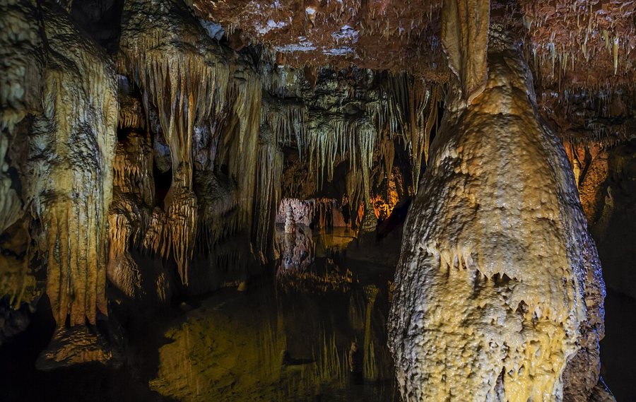 Ein großer heller Stalagmit steht vor Tropfsteinen und einem dunklen unterirdischen Becken in der Höhle Baredine.